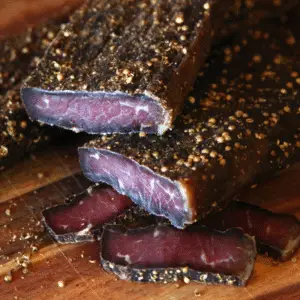 Close-up of traditional South African biltong sliced on a wooden cutting board, seasoned with coriander and black pepper. Dark, cured meat texture with visible spice crust.